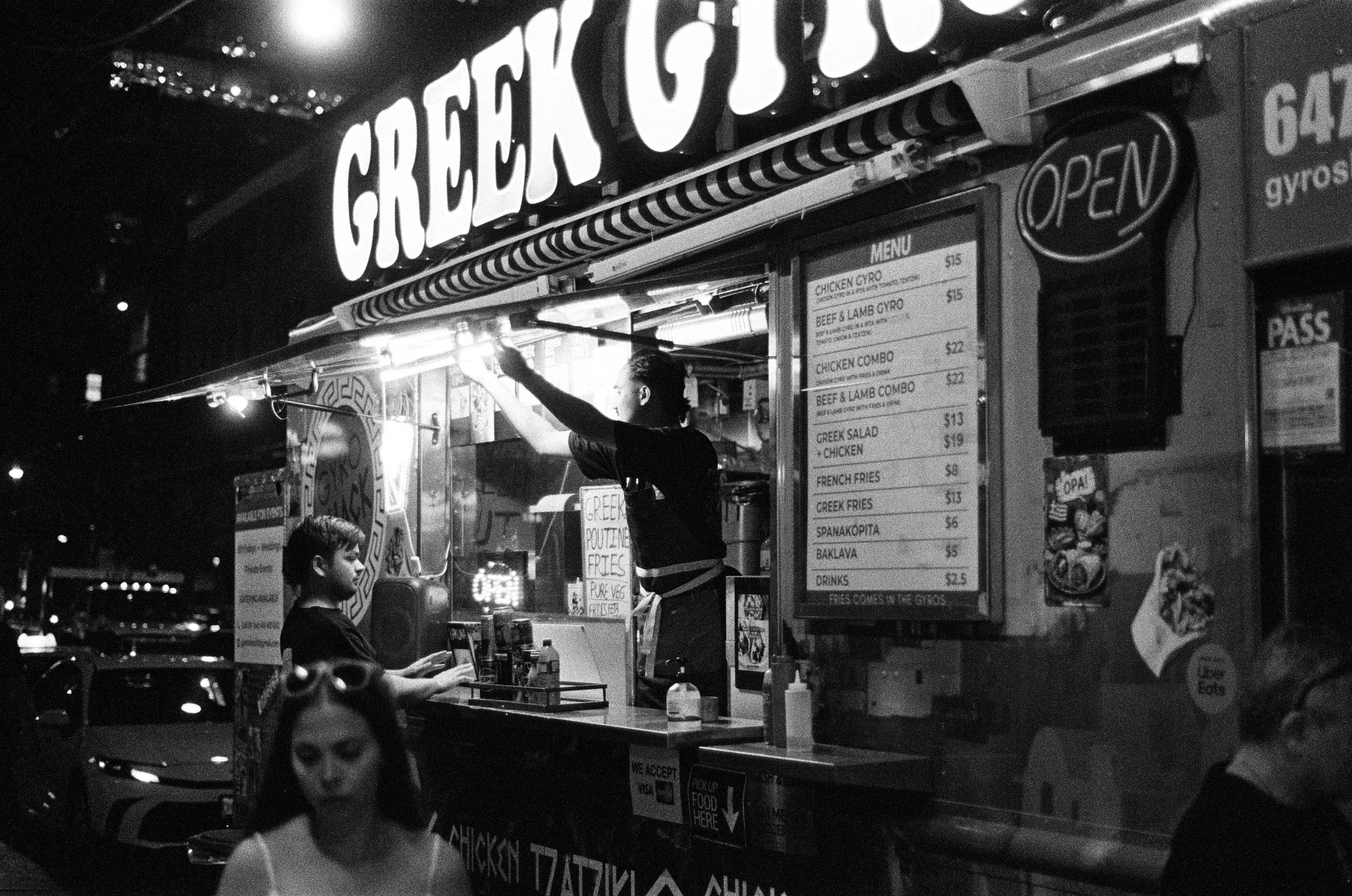 Black-and-white night shot of a food truck under a huge lit GREEK GYRO sign, a cook reaching up at the window