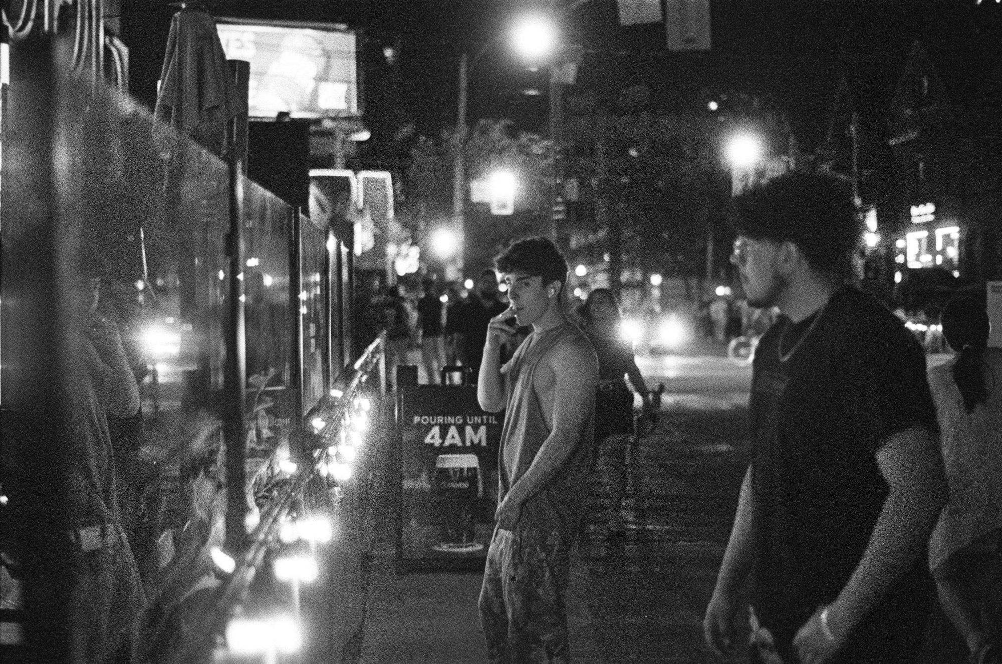 Two young men standing on a sidewalk lit by a bar's lights