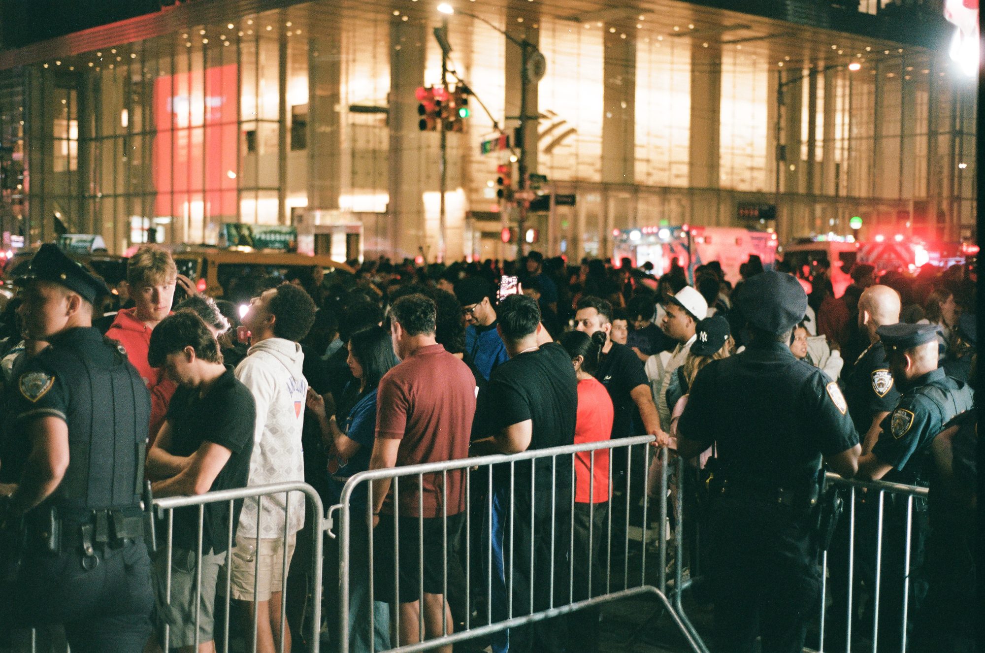 A crowd pressed against police barricades outside a brightly lit arena facade at night