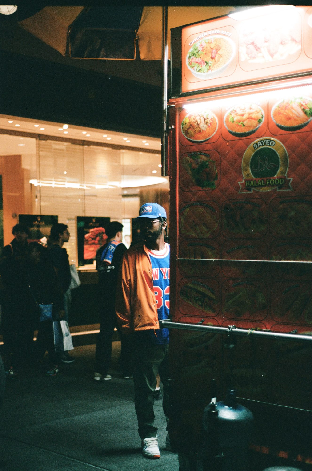 A man in a New York jersey beside a glowing halal food cart