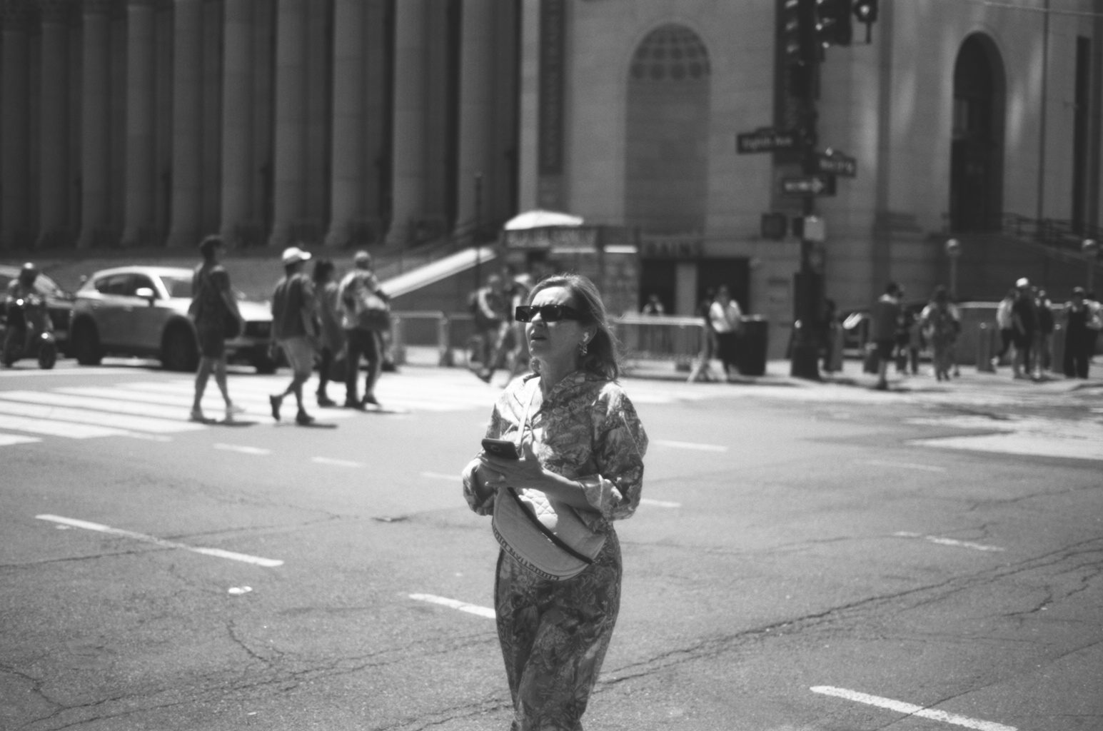 A woman in sunglasses crossing a wide street in hard light