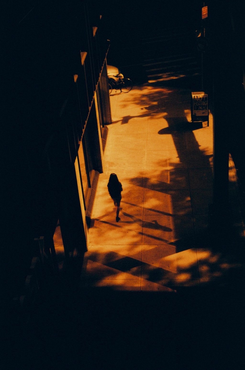Seen from above, a lone figure walks a sunlit pavement in deep red-orange, everything else in shadow